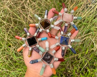 Könnte beinhalten: Sieben Chakra-Kristallheilstäbe, die in einem Kreis angeordnet sind, mit einem pyramidenförmigen Kristall in der Mitte jedes Stabes. Die Kristalle sind in den Farben der Chakren: Rot, Orange, Gelb, Grün, Blau, Indigo und Violett.