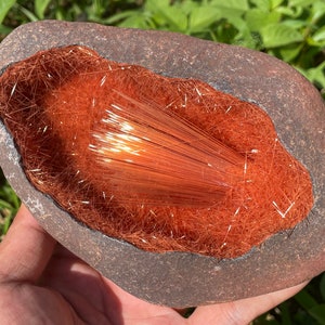 May include: A geode with a natural opening revealing a cluster of orange and red crystals. The crystals are arranged in a fan shape and have a shiny, metallic appearance.