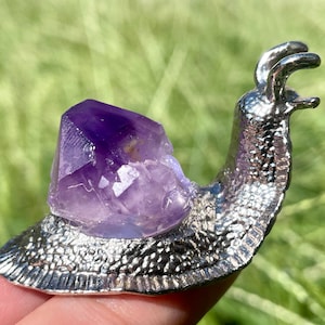 May include: A silver snail figurine with a purple amethyst crystal on its back. The snail is detailed with a textured shell and a raised head.