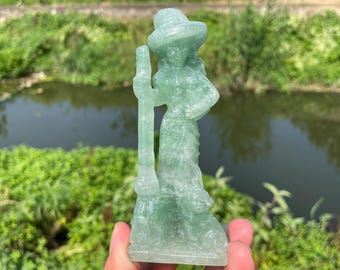 May include: A translucent green figurine of a woman wearing a hat and holding a broom. The statue is carved from a single piece of stone and has a smooth, polished finish. The figurine is a witch, a popular item in the fantasy and Halloween categories.