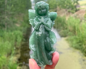 May include: A carved green angel figurine with folded hands and detailed wings. The angel is made of a translucent, jade-like material. The figurine is held in front of a blurred natural background.