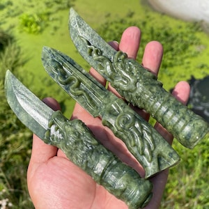 Puede incluir: Tres cuchillos de jade verde con tallas detalladas de dragones y serpientes. Las hojas están pulidas y los mangos presentan diseños intrincados. Los cuchillos se sostienen en una mano.