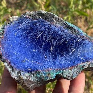 May include: A geode with a blue, fibrous crystal lining. The geode is held in a hand and has a rough, gray exterior.