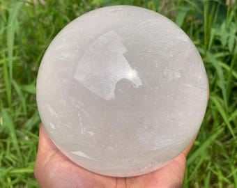May include: A large, clear quartz crystal sphere. The polished surface reflects light, creating a bright, shiny appearance. The sphere is held in a hand, with green grass in the background. This crystal sphere is a decorative item.