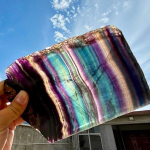 May include: A hand holds a large, rectangular fluorite crystal slab. The stone displays bands of purple, teal, and yellow, with a translucent quality. The background shows a blue sky with scattered clouds, highlighting the crystal's vibrant colors.