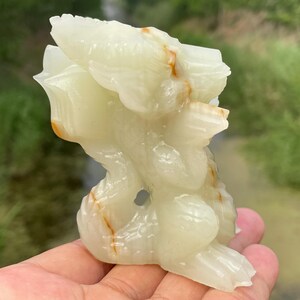May include: A carved onyx sculpture of a mythical creature, featuring a light green color with streaks of orange and brown. The sculpture has a detailed design with rounded shapes and a hole in the center. The background is blurred, with green foliage.