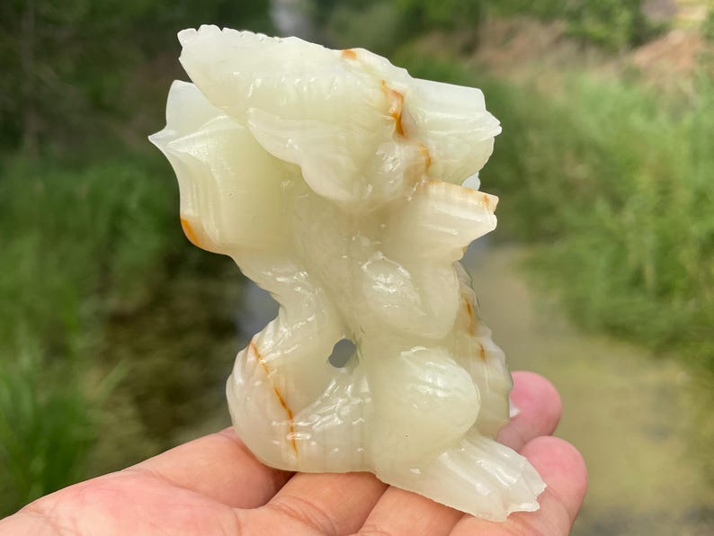 May include: A carved onyx sculpture of a mythical creature, featuring a light green color with streaks of orange and brown. The sculpture has a detailed design with rounded shapes and a hole in the center. The background is blurred, with green foliage.