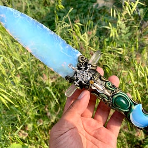 Puede incluir: Una daga de ópalo blanco con un mango verde, dorado y plateado. El mango está decorado con un pentagrama, una luna creciente y una gema verde.