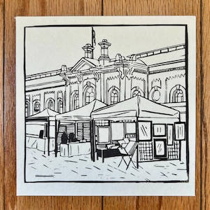 May include: Black and white linocut print depicting an outdoor market scene with vendor tents in front of a large building. The artwork is on a square canvas with a black border, mounted on a wooden surface.