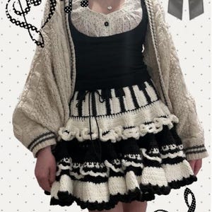 Könnte beinhalten: Ein cremefarbener, übergroßer Cardigan mit schwarzem Besatz wird über einem schwarzen Oberteil und einer weißen Bluse mit schwarzen Punkten getragen. Der Rock ist ein Häkeldesign in Schwarz und Weiß, das Klaviertasten ähnelt. Das Outfit hat ein musikalisches Thema.