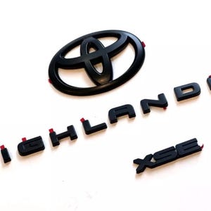 以下が含まれることがあります： 黒いトヨタのエンブレムとハイランダーとXSEの文字。エンブレムは、様式化されたデザインの楕円形です。文字はサンセリフフォントです。文字の裏側には赤い粘着テープが付いています。