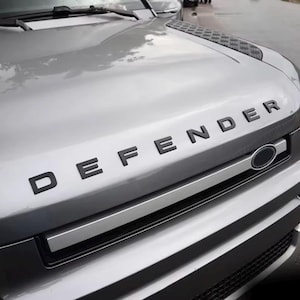Può includere: Un cofano di veicolo color argento con la scritta DEFENDER in lettere nere in grassetto. La parte anteriore include una modanatura orizzontale argento, una sezione della griglia nera e un pannello nero testurizzato sull'angolo superiore.
