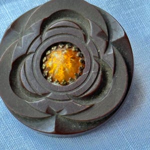 May include: A dark brown, circular vintage button with a floral design. The center features an orange-yellow gem surrounded by small gold-colored beads. The button is set against a light blue fabric background.