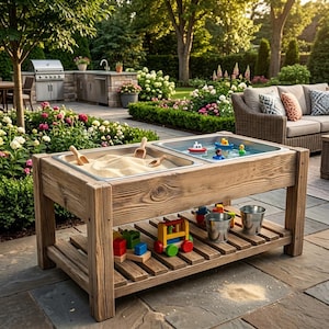 Puede incluir: Una mesa sensorial de madera con dos cubetas rectangulares, una llena de arena y palas de madera, la otra con agua y pequeños barcos de juguete. El estante inferior de listones contiene bloques de madera coloridos, un tren y dos cubos de metal.