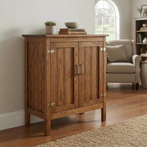 Puede incluir: Un gabinete de madera de color marrón medio con dos puertas de paneles verticales y un diseño de arco apuntado en el panel lateral. El gabinete tiene bisagras y manijas de metal color latón. Una pequeña suculenta y dos libros apilados descansan encima.