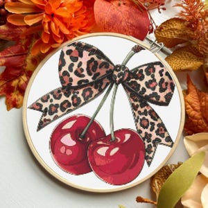 Könnte beinhalten: Ein Kreuzstich-Stickrahmen mit einem Paar roter Kirschen und einer Schleife im Leopardenmuster. Die Kirschen sind tiefrot mit Glanzlichtern, und die Schleife hat ein braun-rosa Muster. Der Rahmen ist vor einem weißen Hintergrund platziert.