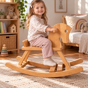 May include: A light-coloured wooden rocking horse with a smooth finish, featuring a curved seat, a stylised head with a circular cutout for an eye, and two curved rockers at the base, designed for a child to sit on and rock back and forth.