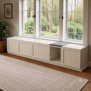 May include: A long, off-white wooden window bench featuring four enclosed storage cabinets with panel detailing, one open cubby space, and a single dark gray rectangular seat cushion positioned on top of the open section beneath a large window.