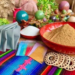 Può includere: Una natura morta con una ciotola rossa piena di polvere marrone, una ciotola bianca con polvere bianca e piccoli pacchetti di pigmenti colorati. Cerchi di legno decorativi e sfere di pietre preziose sono presenti, su un tessuto vibrante e fantasia.