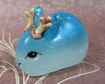 May include: A small, blue, glittery, ceramic figurine of a rabbit with antlers. The rabbit has a white, frosted glaze and a painted black eye.