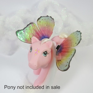 May include: A pink toy pony with large, colourful butterfly wings. The wings display a gradient of colours, including pink, yellow, and green. The pony has blue eyes and a white mane and tail. The text "Pony not included in sale" is at the bottom.