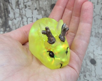 May include: A small, yellow resin figurine of a rabbit with antlers. The rabbit has black eyes and a shiny, iridescent finish.