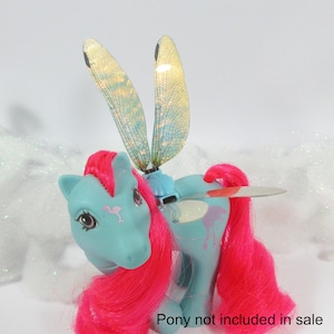 Könnte beinhalten: Ein hellblaues Spielzeugpferd mit einer pinkfarbenen Mähne und einem Schweif, verziert mit schillernden Libellenflügeln. Das Pony hat ein Flamingo-Design auf der Seite und der Wange. Der Text "Pony not included in sale" ist sichtbar.