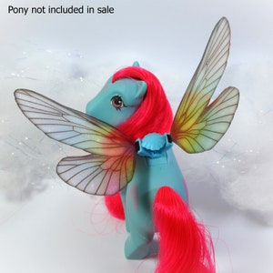 Könnte beinhalten: Ein blaues Spielzeugpferd mit leuchtend pinkfarbenem Haar und Schweif sowie schillernden Feenflügeln. Die Flügel sind mit einem blauen Clip am Rücken des Ponys befestigt. Der Text "Pony not included in sale" ist oben sichtbar.