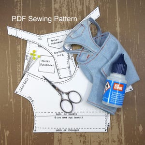 May include: A PDF sewing pattern for a pair of overalls with instructions for cutting and placement of pockets. The pattern is laid out on a wooden surface with a pair of scissors, a bottle of fabric glue, and a pair of blue denim overalls.
