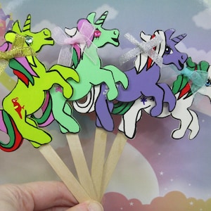Könnte beinhalten: Vier bunte Einhorn-Cupcake-Topper aus Holz. Die Einhörner sind in Grün-, Lila-, Weiß- und Gelbtönen bemalt. Jedes Einhorn hat ein Horn, eine Mähne und einen Schwanz. Die Topper sind an Holzstäbchen befestigt.