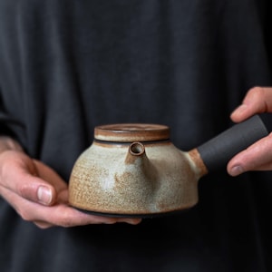 May include: A ceramic teapot with a speckled beige and brown body, a flat lid, and a straight black handle extending from the side. The teapot features a short, centered spout and a textured, earthy finish across its rounded, low-profile base.