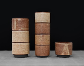 Op de afbeelding: Een set van zeven cilindrische keramische bekers met een gespikkelde beige en bruine afwerking. De bekers zijn gerangschikt in twee verticale stapels van drie en één losse beker, allemaal met een donkerbruine basis en een gladde wand.
