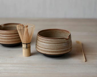 May include: A set of two ceramic bowls with a tan and brown horizontal ribbed texture, a bamboo matcha whisk with thin bristles, and a slender bamboo scoop. The bowls feature a wide opening and a dark base, arranged on a light surface.
