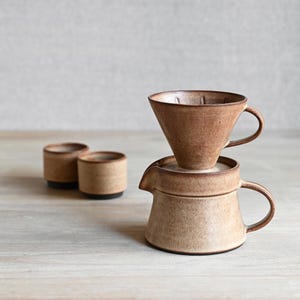 May include: A ceramic coffee pour-over dripper stacked on top of a matching pitcher, accompanied by two small ceramic cups. The set features a textured, speckled tan and brown glaze with a matte finish, showcasing a functional design.