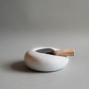 Może przedstawiać: Okrągły, biały ceramiczny uchwyt na kadzidełka o nakrapianej fakturze i gładkiej, zakrzywionej krawędzi. Kawałek jasnego drewna z zwęglonym, czarnym końcem spoczywa na otworze uchwytu, wystając nieco poza jego krawędź.