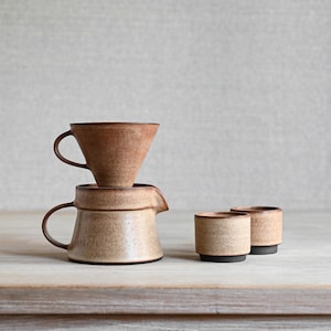 May include: A ceramic coffee pour-over dripper stacked on a matching creamer pitcher, accompanied by two small cylindrical cups. The set features a speckled, earthy brown and beige glaze with a smooth, matte finish and dark brown base accents.