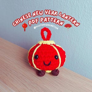 Può includere: Una lanterna cinese del Capodanno in crochet rosso e giallo con un viso sorridente. La lanterna ha un anello in cima per appenderla. Il testo "CHINESE NEW YEAR LANTERN PDF PATTERN @CUSTOMCROCHETSSG" è sopra la lanterna.