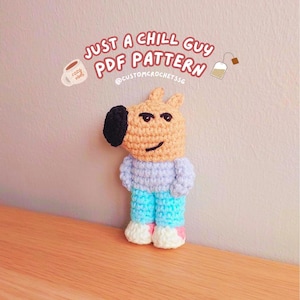 Op de afbeelding: Een gehaakt amigurumi-speelgoed van een cartoonhond die een lichtblauw shirt en een blauwe broek draagt. Het speelgoed staat op een houten oppervlak. De tekst "JUST A CHILL GUY PDF PATTERN @CUSTOMCROCHETSS" staat boven het speelgoed.