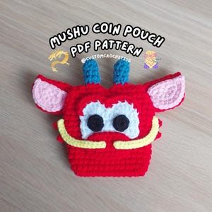 May include: A red crocheted coin pouch in the shape of a cartoon character with the text "Mushu Coin Pouch PDF Pattern". The pouch has blue horns, pink ears, and yellow accents. Black eyes and a white face complete the design.