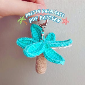 May include: A teal blue crocheted palm tree keychain with a brown trunk and a silver metal ring. The keychain is labeled "Pretty Palm Tree PDF Pattern" and "@customcrochetissg".