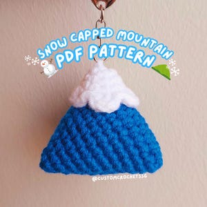 May include: A blue crocheted mountain keychain with a white crocheted snow cap. The keychain has a silver ring and is labelled "Snow Capped Mountain PDF Pattern".