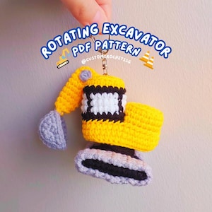 Op de afbeelding: Een geel gehaakt speelgoed-graafmachine met een grijze bak en zwarte rupsbanden. Het speelgoed is bevestigd aan een sleutelhanger met een zilveren ring. De tekst "Rotating Excavator PDF Pattern @customcrochetissg" is in blauw en wit op een witte achtergrond geschreven.