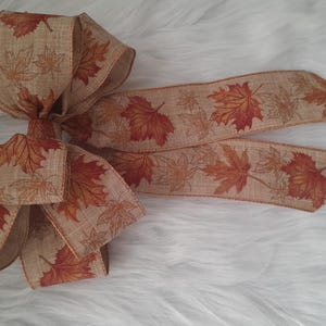 May include: A decorative bow made of tan ribbon, featuring a repeating pattern of autumn leaves in shades of orange and brown. The bow is ideal for fall-themed crafts and decorations.