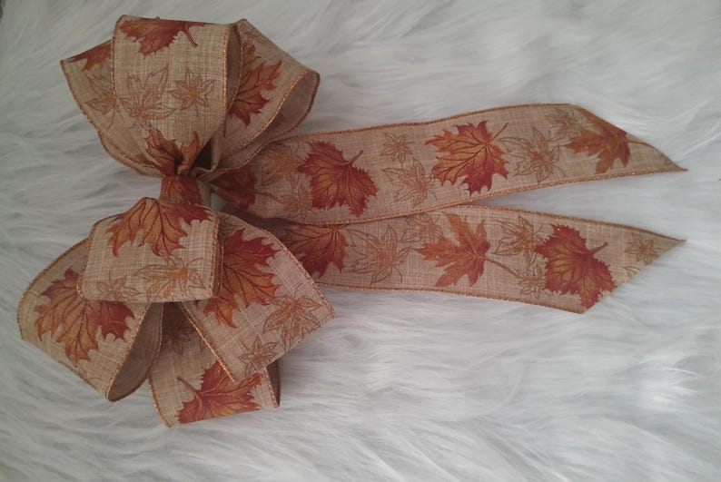 May include: A decorative bow made of tan ribbon, featuring a repeating pattern of autumn leaves in shades of orange and brown. The bow is ideal for fall-themed crafts and decorations.