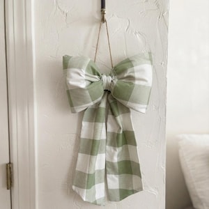 May include: A decorative fabric bow featuring a sage green and white buffalo check pattern. The bow is suspended by a thin twine string from a vintage-style metal key hook. It has two large loops and two long, rectangular tails hanging downward.