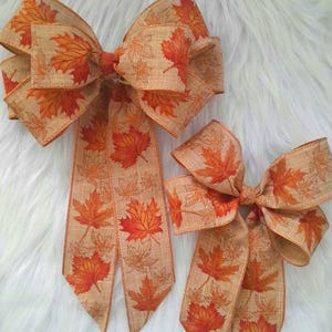 May include: Two decorative fall-themed bows. The bows are made of tan ribbon with a repeating pattern of orange and red maple leaves. The larger bow is approximately 10 inches wide, and the smaller bow is about 6 inches wide. The bows are set against a white, fluffy background.