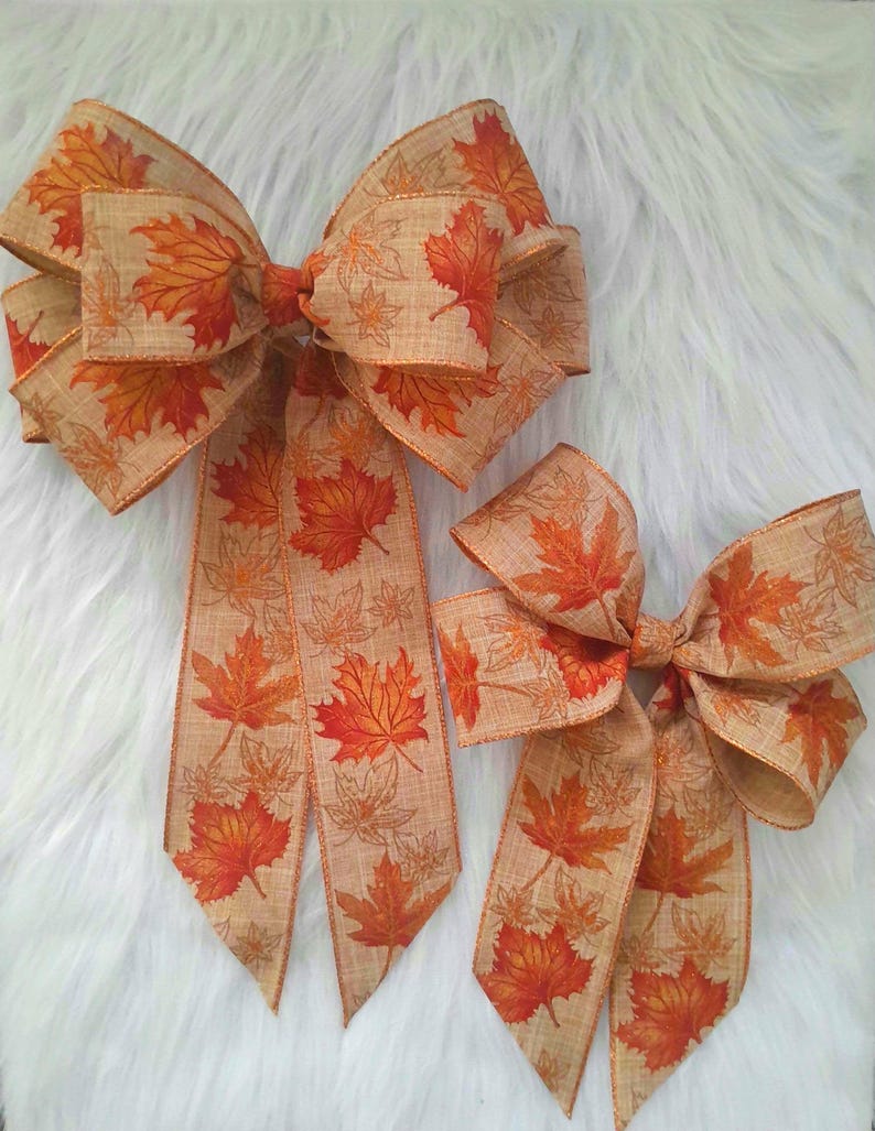May include: Two decorative fall-themed bows. The bows are made of tan ribbon with a repeating pattern of orange and red maple leaves. The larger bow is approximately 10 inches wide, and the smaller bow is about 6 inches wide. The bows are set against a white, fluffy background.