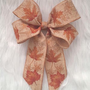 May include: A decorative bow featuring a tan ribbon with a fall leaf pattern in shades of orange and brown. The bow has multiple loops and long tails, perfect for autumn-themed crafts and decorations.