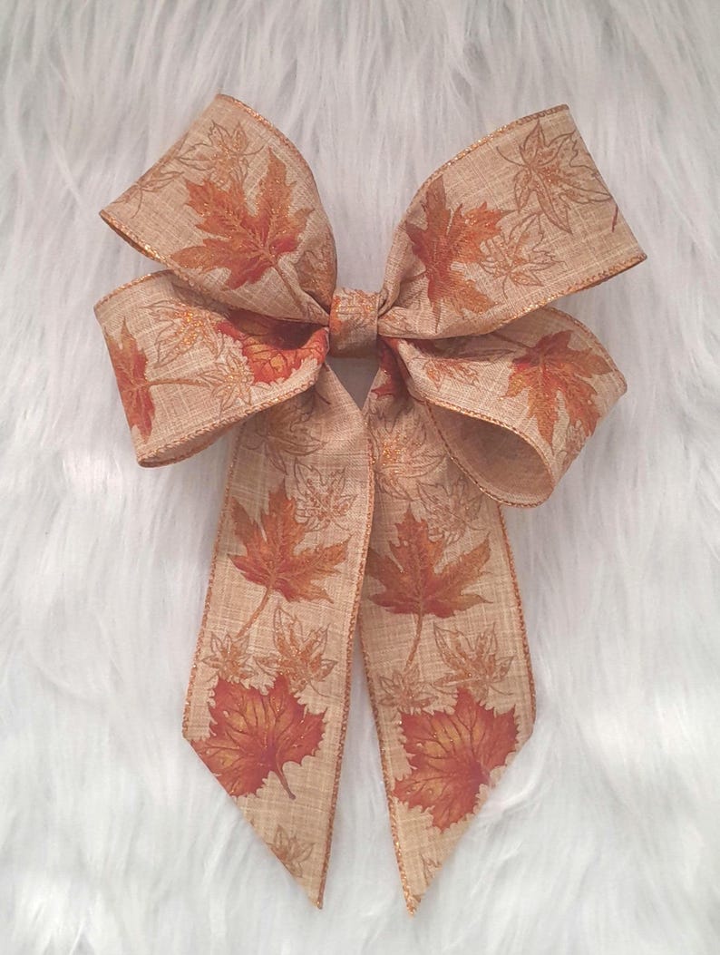 May include: A decorative bow featuring a tan ribbon with a fall leaf pattern in shades of orange and brown. The bow has multiple loops and long tails, perfect for autumn-themed crafts and decorations.