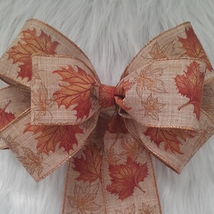May include: A decorative bow featuring a tan fabric with a leaf pattern in shades of orange and red. The bow has a gold glitter trim and is tied in the center. The bow is likely used for seasonal decor, such as autumn or fall.